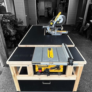 K&ouml;nnte beinhalten: Eine ma&szlig;gefertigte Holzbearbeitungs-Werkbank mit schwarzer Oberseite und einer gelb-grauen DeWalt Tischkreiss&auml;ge und Gehrungss&auml;ge. Die Werkbank hat einen Holzrahmen und eine schwarze Schublade. Die Werkbank steht auf Rollen.