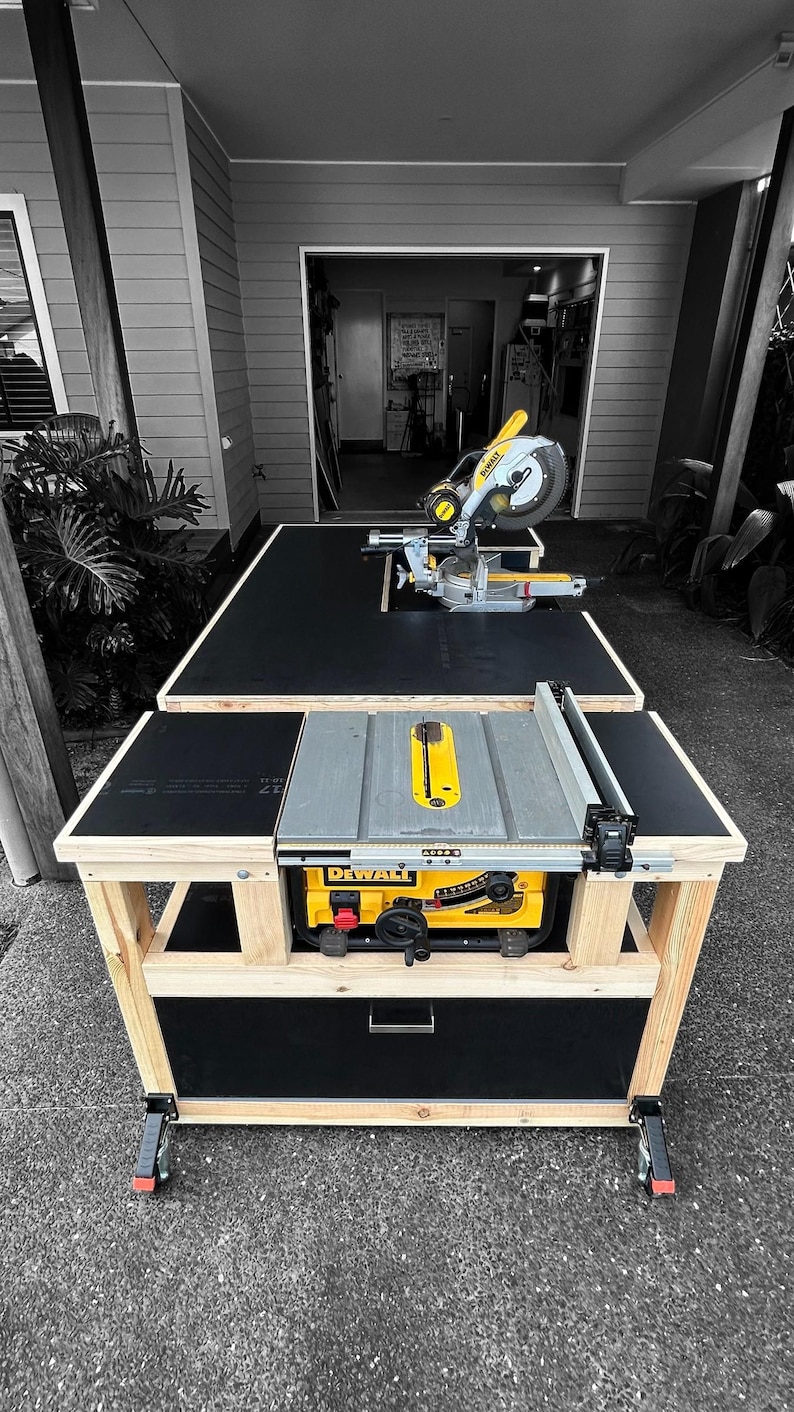 K&ouml;nnte beinhalten: Eine ma&szlig;gefertigte Holzbearbeitungs-Werkbank mit schwarzer Oberseite und einer gelb-grauen DeWalt Tischkreiss&auml;ge und Gehrungss&auml;ge. Die Werkbank hat einen Holzrahmen und eine schwarze Schublade. Die Werkbank steht auf Rollen.