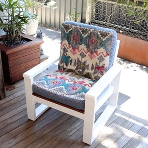 May include: White wooden outdoor chair with a patterned cushion. The cushion features a design with teal, pink, and cream colors. The chair is on a wooden deck, with a wooden planter and a bench in the background.