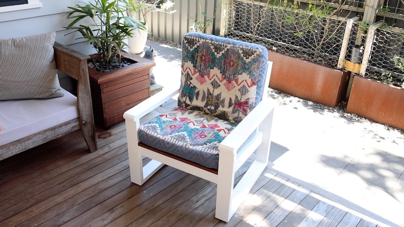 May include: White wooden outdoor chair with a patterned cushion. The cushion features a design with teal, pink, and cream colors. The chair is on a wooden deck, with a wooden planter and a bench in the background.