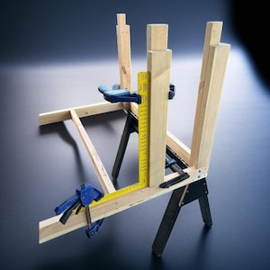 K&ouml;nnte beinhalten: Ein Holzbearbeitungsprojekt in Arbeit, bei dem Holzbalken mit blauen Klemmen zusammengeklemmt werden. Ein gelbes Lineal wird zum Messen verwendet. Die Struktur wird von schwarzen S&auml;geb&ouml;cken getragen, was ein DIY- oder Konstruktionsthema zeigt.