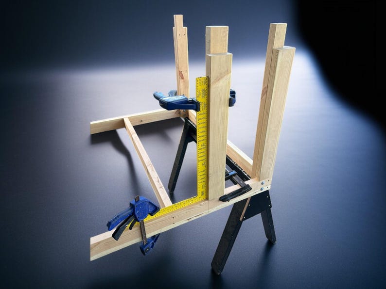 K&ouml;nnte beinhalten: Ein Holzbearbeitungsprojekt in Arbeit, bei dem Holzbalken mit blauen Klemmen zusammengeklemmt werden. Ein gelbes Lineal wird zum Messen verwendet. Die Struktur wird von schwarzen S&auml;geb&ouml;cken getragen, was ein DIY- oder Konstruktionsthema zeigt.