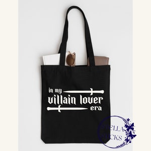 Pode incluir: Sacola tote preta com texto branco que diz "in my villain lover era" com duas espadas acima e abaixo do texto.