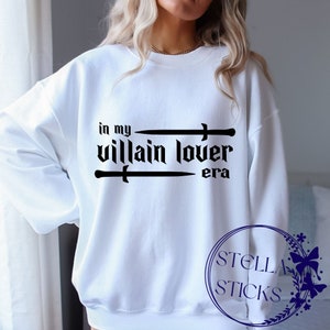 Pode incluir: Camisola branca com texto preto que diz "in my villain lover era" com duas espadas cruzadas acima do texto.
