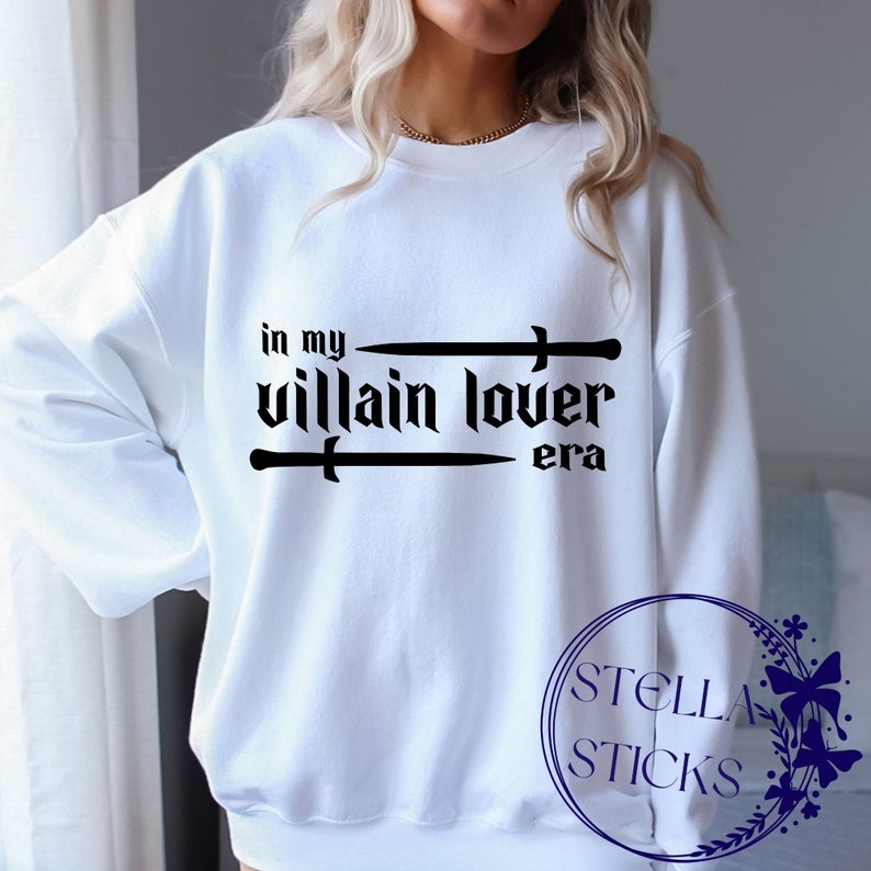 Pode incluir: Camisola branca com texto preto que diz "in my villain lover era" com duas espadas cruzadas acima do texto.