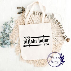 Pode incluir: Sacola de lona branca com texto preto que diz "in my villain lover era" com duas espadas cruzadas acima do texto. A sacola tem al&ccedil;as pretas e est&aacute; sobre uma superf&iacute;cie branca com uma tape&ccedil;aria de macram&ecirc; tecida no fundo.