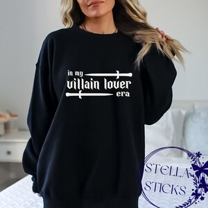 Pode incluir: Camisola de moletom preta com texto branco que diz "in my villain lover era" com duas espadas acima e abaixo do texto.