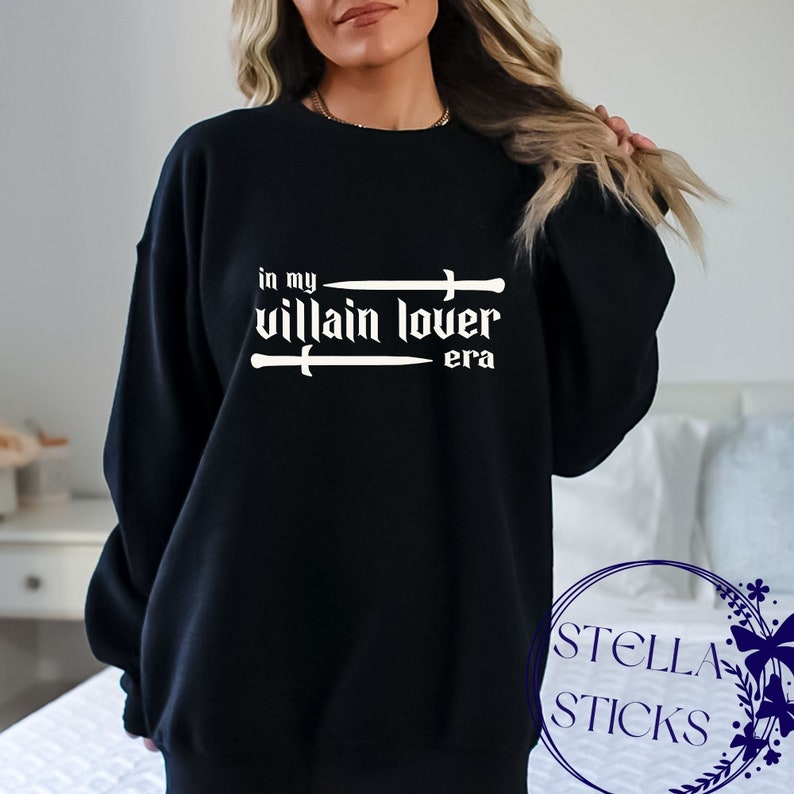 Pode incluir: Camisola de moletom preta com texto branco que diz "in my villain lover era" com duas espadas acima e abaixo do texto.