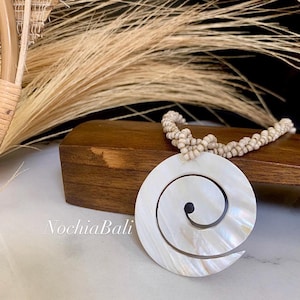 Puede incluir: Un collar de conchas blancas con un gran colgante de concha blanca en forma de espiral. El collar está hecho de pequeñas cuentas blancas y se muestra sobre una superficie de madera.