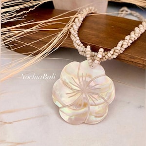 May include: A pearlescent flower pendant necklace with a carved floral design. The pendant hangs from a multi-strand beaded necklace. The necklace is displayed on a wooden surface with dried grass in the background. The text "NochiaBali" is visible.
