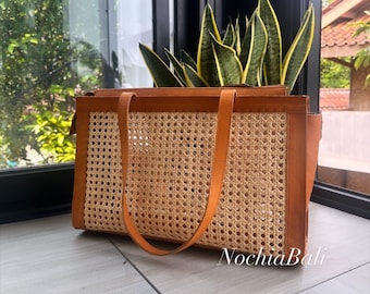 May include: A brown leather tote bag with a woven rattan panel. The bag has two leather straps and is sitting on a tiled surface.