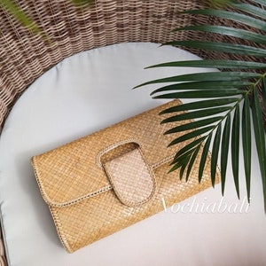 Könnte beinhalten: Eine Clutch aus geflochtenem Stroh in Beige mit rechteckiger Form und Klappenverschluss. Die Clutch hat ein gewebtes Muster. Die Clutch liegt auf einem weißen Kissen, mit grünen Blättern im Hintergrund. Der Text "Vochiabali" ist sichtbar.