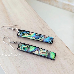 May include: A pair of rectangular earrings with iridescent abalone shell inlays. The earrings have a black border and silver-colored ear wires. The text "Nochiabali" is visible on the wooden surface.