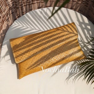 May include: A woven clutch bag in a light brown color. The bag has a flap closure and is made of a natural material. The bag is sitting on a white surface with a shadow of a palm leaf.