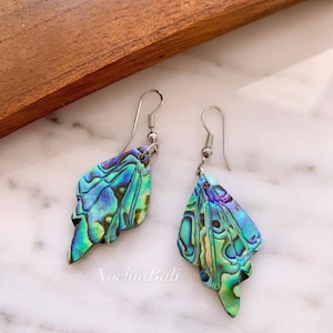 Könnte beinhalten: Ein Paar Ohrringe aus irisierendem grün-blauem Abalone-Muschelschalen mit silbernen Haken. Die Ohrringe sind tropfenförmig und haben ein mehrschichtiges Design.