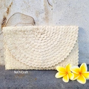 K&ouml;nnte beinhalten: Eine wei&szlig;e geflochtene Clutch mit einem abgerundeten Klappdeckel. Die Clutch ist aus einem strukturierten Material gefertigt und hat einen dezenten Glanz. Die Clutch liegt auf einer grauen Oberfl&auml;che mit zwei wei&szlig;en Frangipani-Bl&uuml;ten.