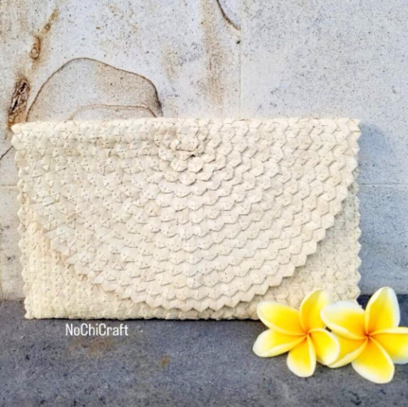 K&ouml;nnte beinhalten: Eine wei&szlig;e geflochtene Clutch mit einem abgerundeten Klappdeckel. Die Clutch ist aus einem strukturierten Material gefertigt und hat einen dezenten Glanz. Die Clutch liegt auf einer grauen Oberfl&auml;che mit zwei wei&szlig;en Frangipani-Bl&uuml;ten.