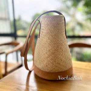 Puede incluir: Un bolso tote de paja tejido con correas y ribetes de cuero marr&oacute;n. El bolso tiene un fondo redondeado y una abertura ancha. El bolso est&aacute; sobre una superficie de madera.