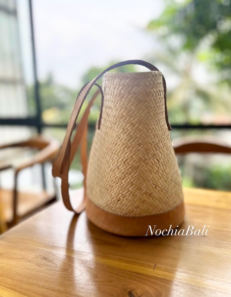 Puede incluir: Un bolso tote de paja tejido con correas y ribetes de cuero marr&oacute;n. El bolso tiene un fondo redondeado y una abertura ancha. El bolso est&aacute; sobre una superficie de madera.