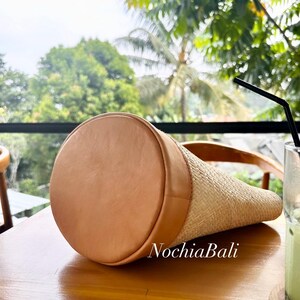 Puede incluir: Un bolso tote de paja trenzada con ribete de cuero marr&oacute;n y el texto "NochiaBali" en el lateral.