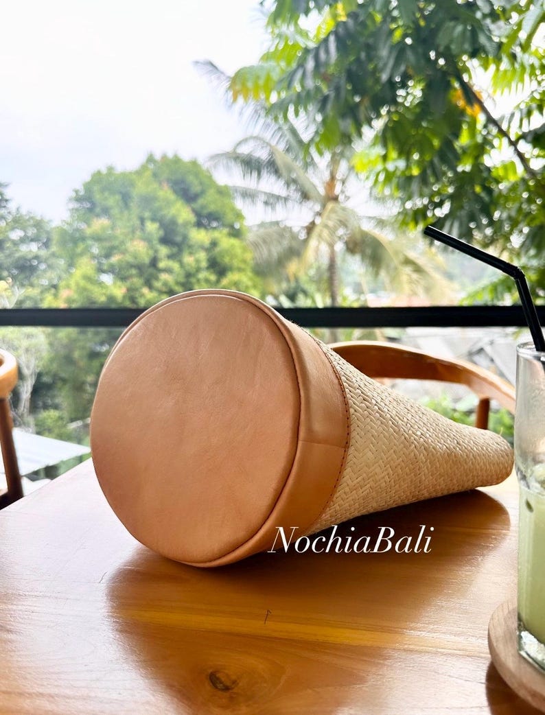 Puede incluir: Un bolso tote de paja trenzada con ribete de cuero marr&oacute;n y el texto "NochiaBali" en el lateral.