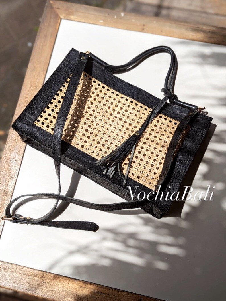 May include: A black leather tote bag with a woven rattan panel and a long tassel. The bag has a top handle and a long shoulder strap. The bag is made by Nochia Bali.