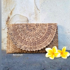 K&ouml;nnte beinhalten: Eine gewebte Clutch-Tasche aus nat&uuml;rlichen Materialien, in brauner Farbe und mit einem einzigartigen Muster. Die Tasche liegt auf einer strukturierten Oberfl&auml;che mit zwei gelben Blumen im Vordergrund. Der Text "NoChiCraft" ist in der linken unteren Ecke des Bildes sichtbar.