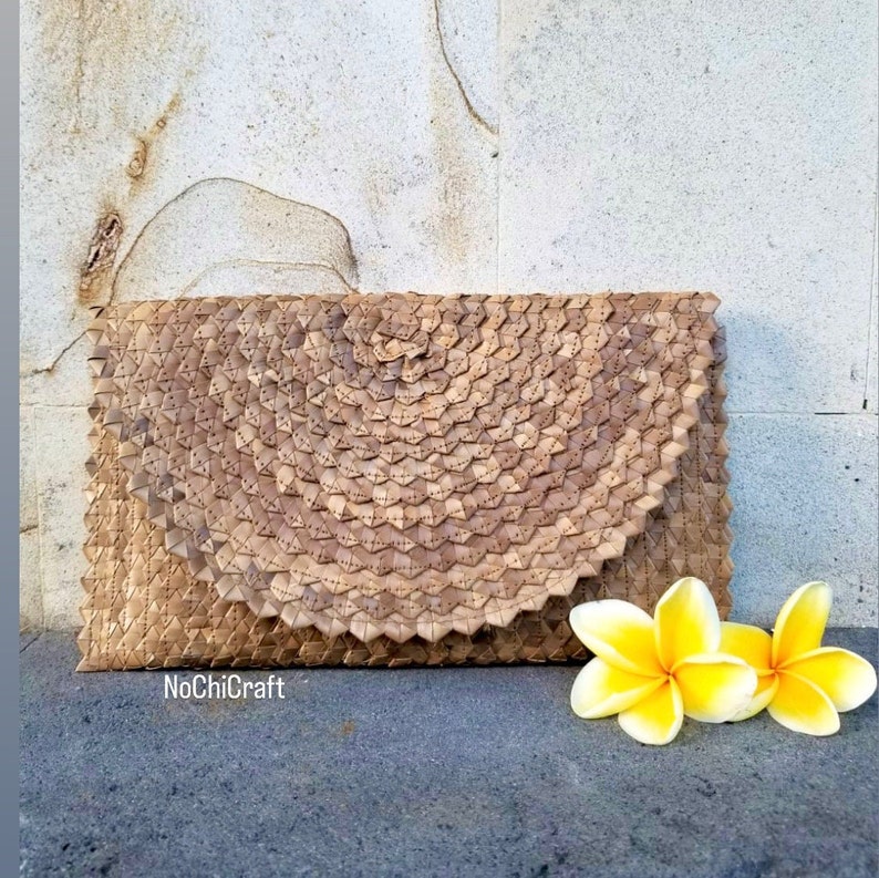 K&ouml;nnte beinhalten: Eine gewebte Clutch-Tasche aus nat&uuml;rlichen Materialien, in brauner Farbe und mit einem einzigartigen Muster. Die Tasche liegt auf einer strukturierten Oberfl&auml;che mit zwei gelben Blumen im Vordergrund. Der Text "NoChiCraft" ist in der linken unteren Ecke des Bildes sichtbar.