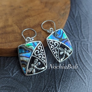 May include: Silver dangle earrings with a square-shaped abalone shell inlay and a dragonfly design. The earrings have a silver hook fastening.