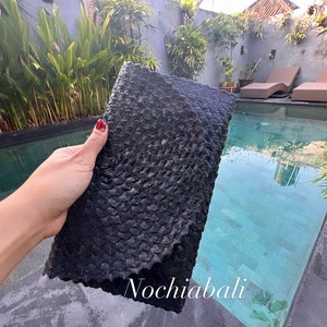 K&ouml;nnte beinhalten: Schwarze geflochtene Clutch mit einzigartigem, strukturiertem Design. Die Clutch wird von einer Person in der Hand gehalten, in der N&auml;he eines Schwimmbeckens.