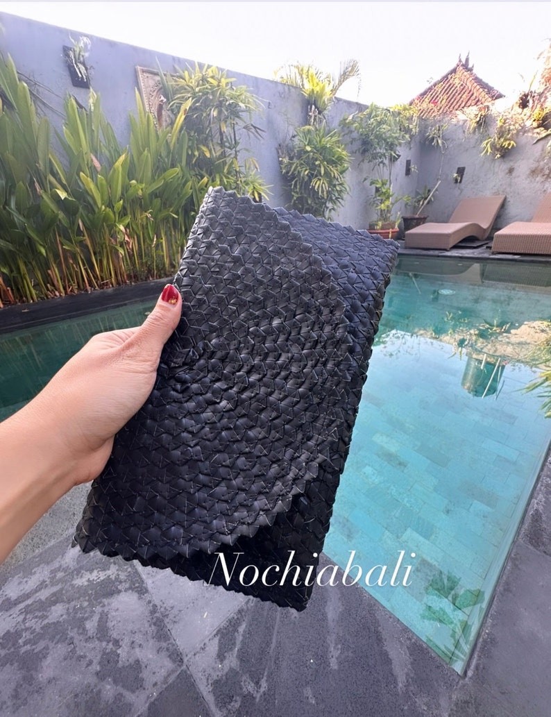 K&ouml;nnte beinhalten: Schwarze geflochtene Clutch mit einzigartigem, strukturiertem Design. Die Clutch wird von einer Person in der Hand gehalten, in der N&auml;he eines Schwimmbeckens.