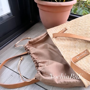 Puede incluir: Un bolso tote de paja tejido con una correa de cuero marr&oacute;n y un forro de tela marr&oacute;n. El bolso est&aacute; abierto y el forro es visible. El bolso est&aacute; sobre una superficie de madera.