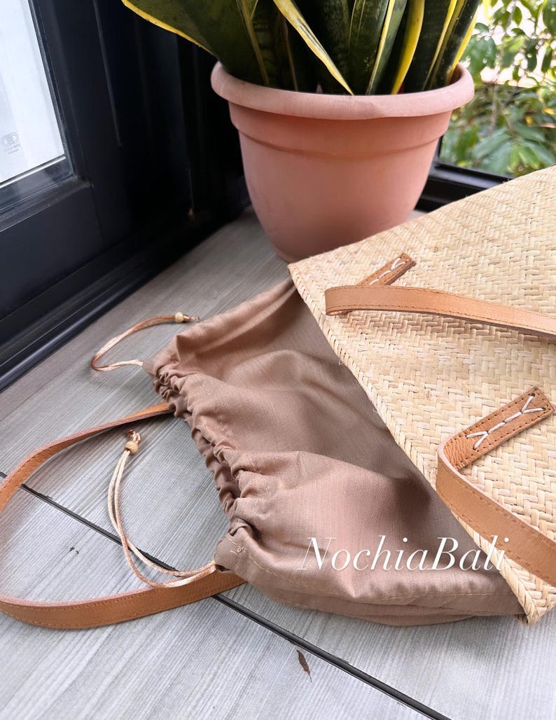 Puede incluir: Un bolso tote de paja tejido con una correa de cuero marr&oacute;n y un forro de tela marr&oacute;n. El bolso est&aacute; abierto y el forro es visible. El bolso est&aacute; sobre una superficie de madera.