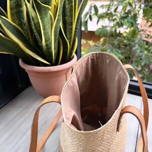 Puede incluir: Un bolso tote tejido de color marr&oacute;n claro con correas de cuero. El bolso est&aacute; abierto y el interior est&aacute; forrado con un tejido marr&oacute;n claro. El bolso est&aacute; sobre una superficie de madera.