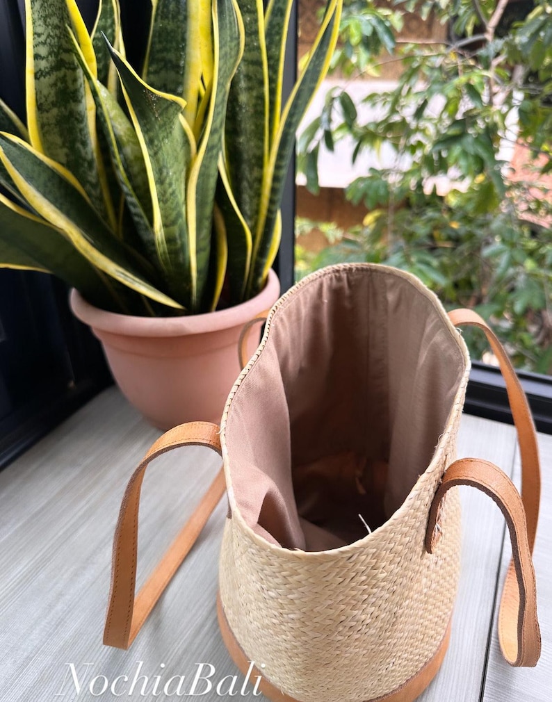 Puede incluir: Un bolso tote tejido de color marr&oacute;n claro con correas de cuero. El bolso est&aacute; abierto y el interior est&aacute; forrado con un tejido marr&oacute;n claro. El bolso est&aacute; sobre una superficie de madera.