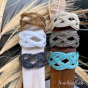 Puede incluir: Seis pulseras de cuentas en diferentes colores, incluyendo blanco, dorado, plateado, turquesa y azul iridiscente. Las pulseras se muestran en soportes de madera. El texto "Nocha Bali" es visible en la parte inferior de la imagen.