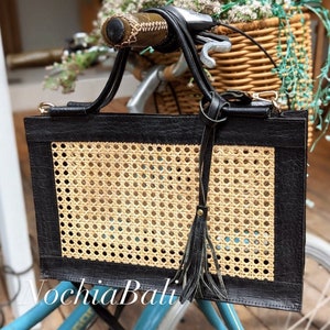 May include: A black leather tote bag with a woven rattan panel and a black leather tassel. The bag is hanging on the handlebars of a blue bicycle.