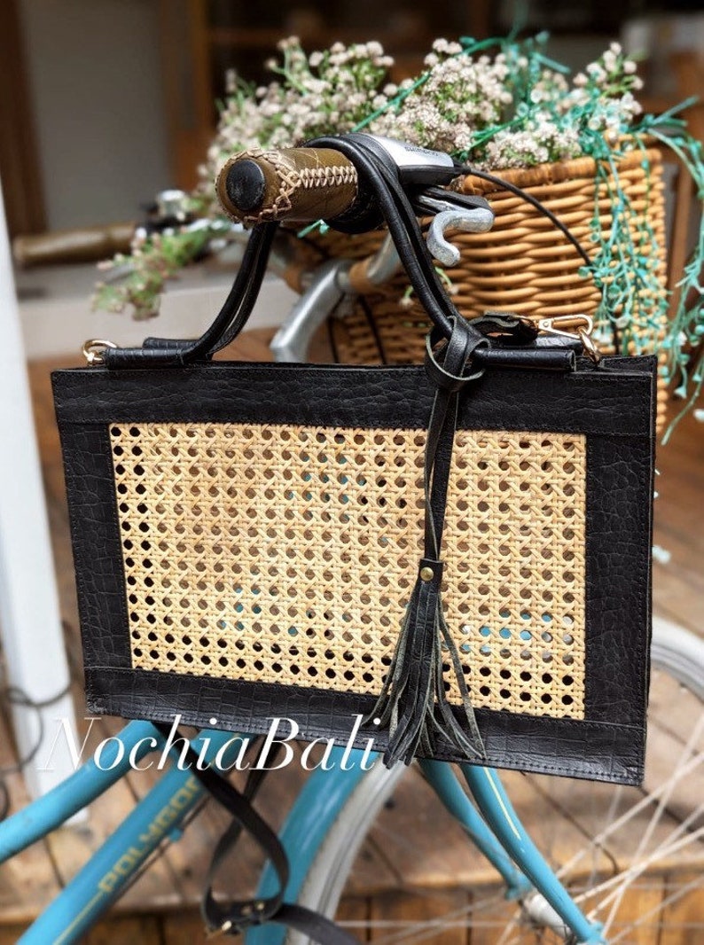May include: A black leather tote bag with a woven rattan panel and a black leather tassel. The bag is hanging on the handlebars of a blue bicycle.