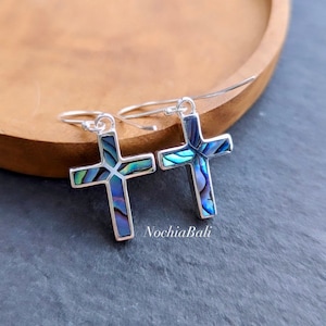 May include: A pair of silver hoop earrings with a blue and green abalone shell cross pendant. The cross is made of silver and has a shiny finish.