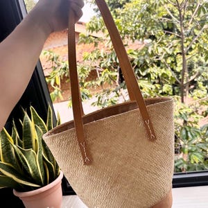 Puede incluir: Un bolso tote de paja tejido con asas y ribetes de cuero marr&oacute;n. El bolso tiene forma cil&iacute;ndrica y est&aacute; hecho con un patr&oacute;n tejido.