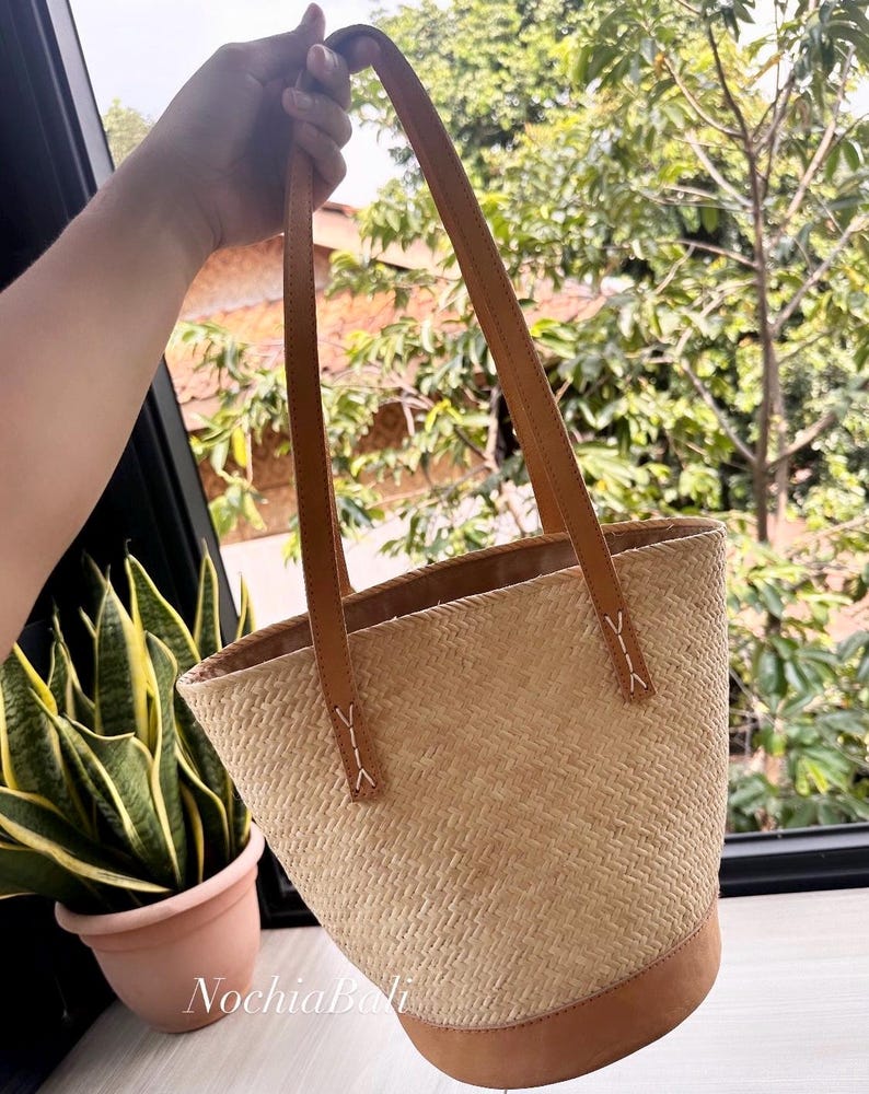 Puede incluir: Un bolso tote de paja tejido con asas y ribetes de cuero marr&oacute;n. El bolso tiene forma cil&iacute;ndrica y est&aacute; hecho con un patr&oacute;n tejido.
