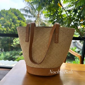 Puede incluir: Un bolso tote tejido de color marr&oacute;n claro con asas y ribetes de cuero. El bolso est&aacute; sobre una mesa de madera.