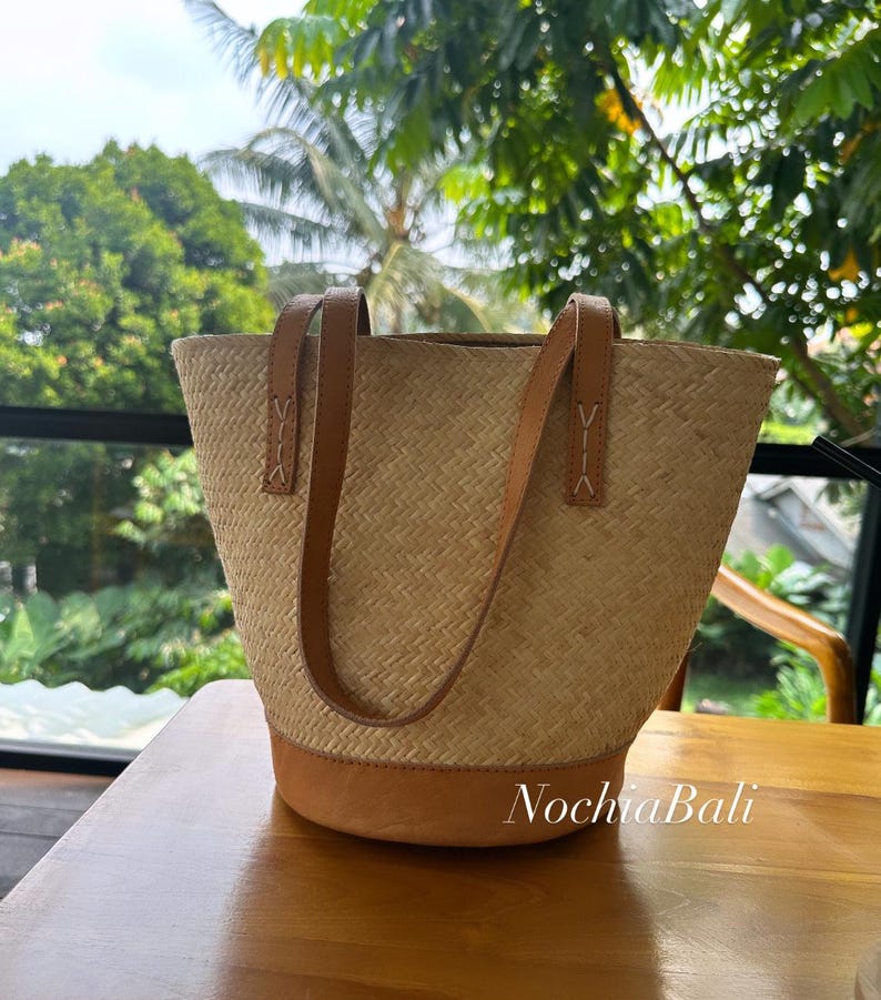 Puede incluir: Un bolso tote tejido de color marr&oacute;n claro con asas y ribetes de cuero. El bolso est&aacute; sobre una mesa de madera.