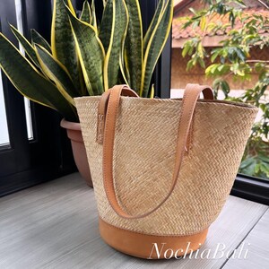 Puede incluir: Un bolso tote tejido de color beige con correas de cuero marr&oacute;n y una base de cuero marr&oacute;n. El bolso est&aacute; sobre una superficie de madera.