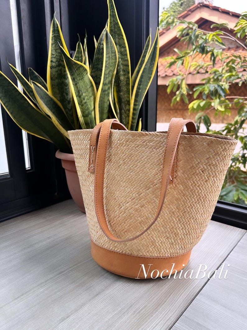 Puede incluir: Un bolso tote tejido de color beige con correas de cuero marr&oacute;n y una base de cuero marr&oacute;n. El bolso est&aacute; sobre una superficie de madera.