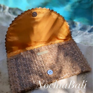 K&ouml;nnte beinhalten: Eine geflochtene braune Lederclutch mit einem goldenen Knopfverschluss und einem gelben Satin-Futter. Die Clutch hat einen gewellten Rand und der Markenname "Nochla Bali" ist unten sichtbar.
