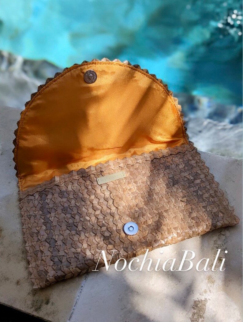 K&ouml;nnte beinhalten: Eine geflochtene braune Lederclutch mit einem goldenen Knopfverschluss und einem gelben Satin-Futter. Die Clutch hat einen gewellten Rand und der Markenname "Nochla Bali" ist unten sichtbar.