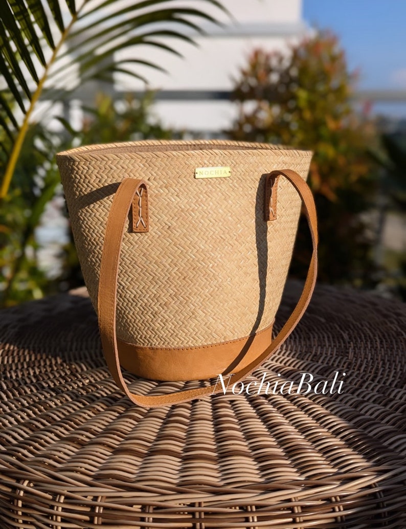 Puede incluir: Un bolso tote de paja tejido con asas de cuero y fondo de cuero. El bolso tiene una etiqueta dorada que dice "Nochhia". El bolso est&aacute; sobre una superficie de mimbre tejido.