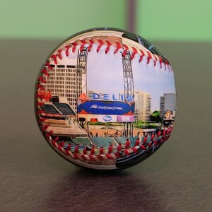 May include: A white baseball with red stitching and a colorful image of a baseball stadium with the text "DELUXE" and "SunTrust Park" printed on it.