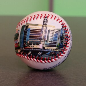 May include: White baseball with red stitching and a printed image of a city skyline. The skyline is a mix of blue, gray, and white buildings.